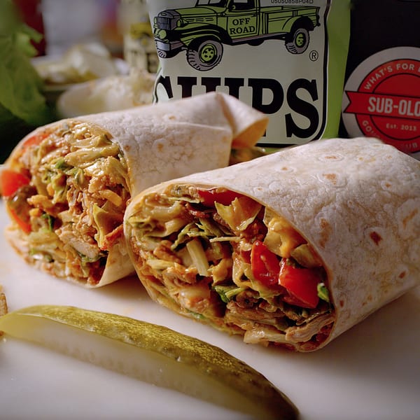 Two Sub-Ology wraps filled with chicken, lettuce, and tomatoes, served with a pickle spear.