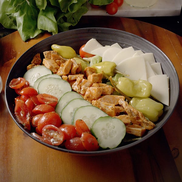 A Sub-Ology salad with chicken, cucumbers, tomatoes, cheese, and peppers in a dark bowl.
