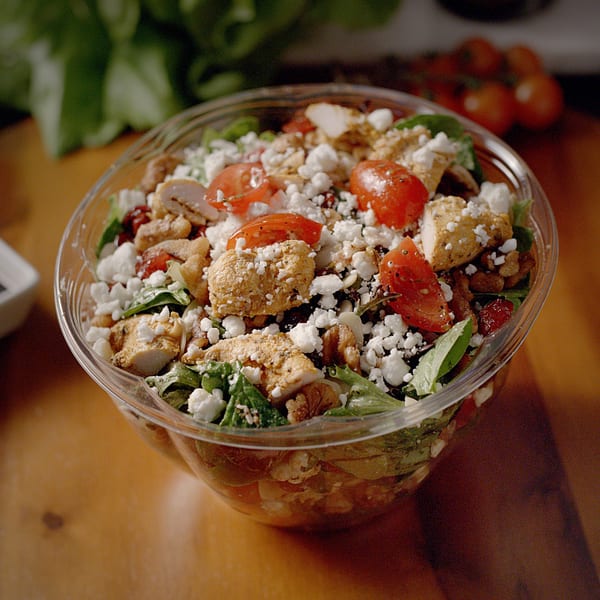 A Sub-Ology salad with chicken, tomatoes, feta cheese, and greens in a clear plastic bowl.