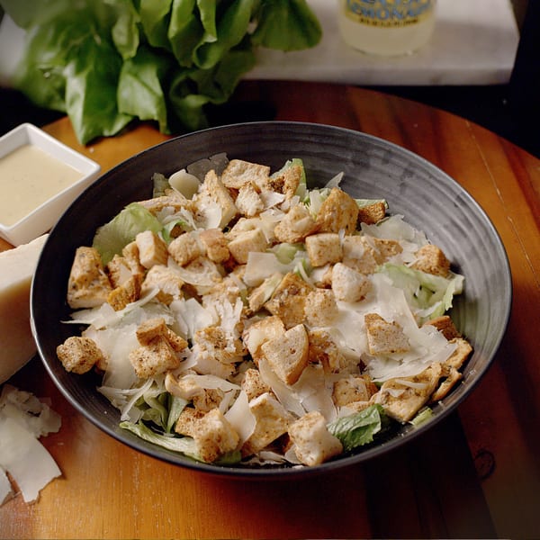 A Caesar salad with lettuce, croutons, and shaved Parmesan cheese in a dark bowl.