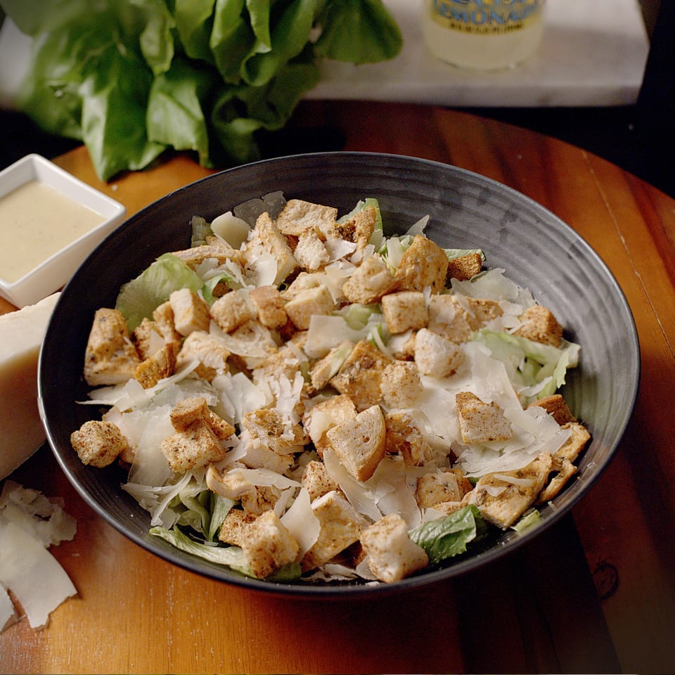 A Caesar salad with lettuce, croutons, and shaved Parmesan cheese in a dark bowl.