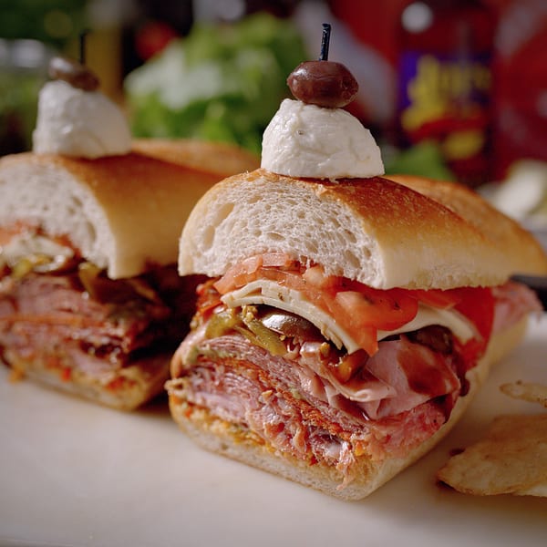 Two Sub-Ology sandwiches, cut in half, overflowing with meats, cheeses, and vegetables, on a white surface.