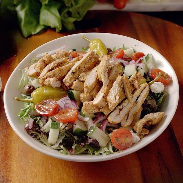 A white bowl of salad with sliced chicken, tomatoes, cucumbers, red onion, and a pepperoncini.