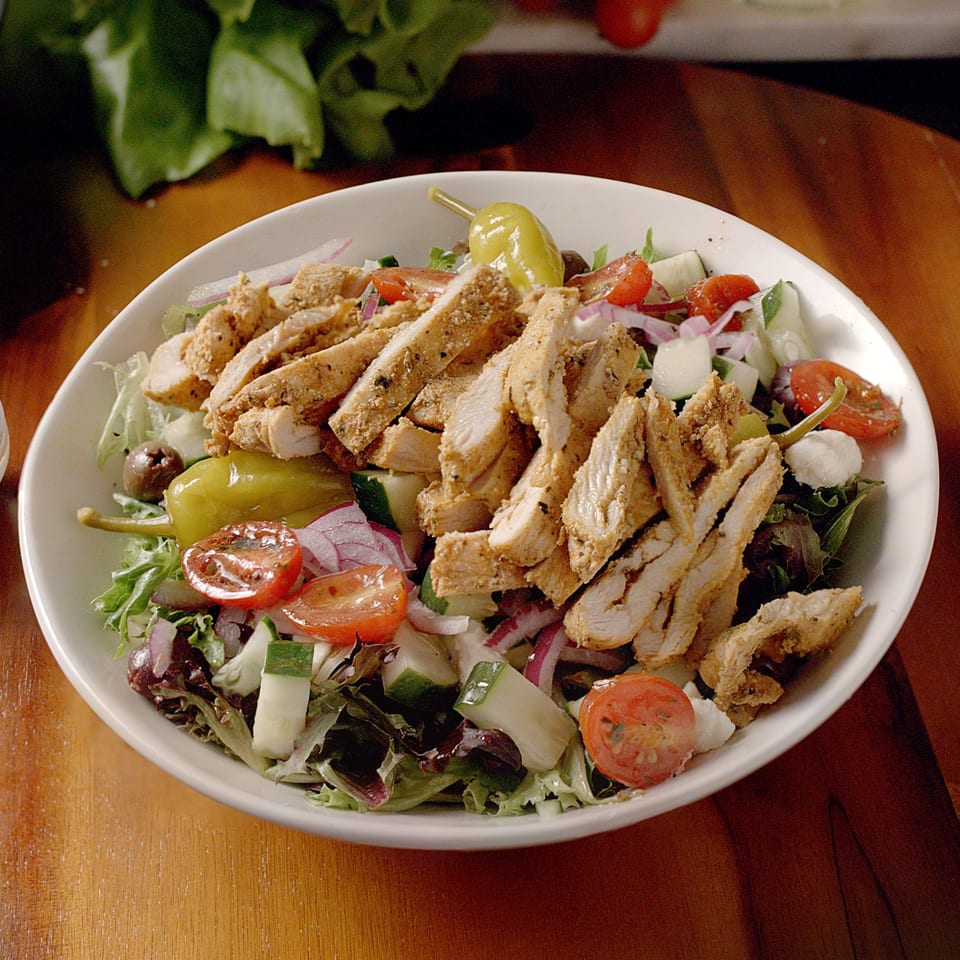 A white bowl of salad with sliced chicken, tomatoes, cucumbers, red onion, and a pepperoncini.